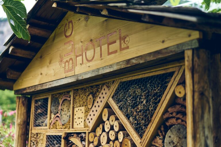 Een houten constructie met het label "Bug Hotel" herbergt verschillende compartimenten gevuld met natuurlijke materialen zoals hout, bamboe en bladeren, ontworpen om insecten onderdak te bieden. Het dak is schuin en groen is zichtbaar op de achtergrond.