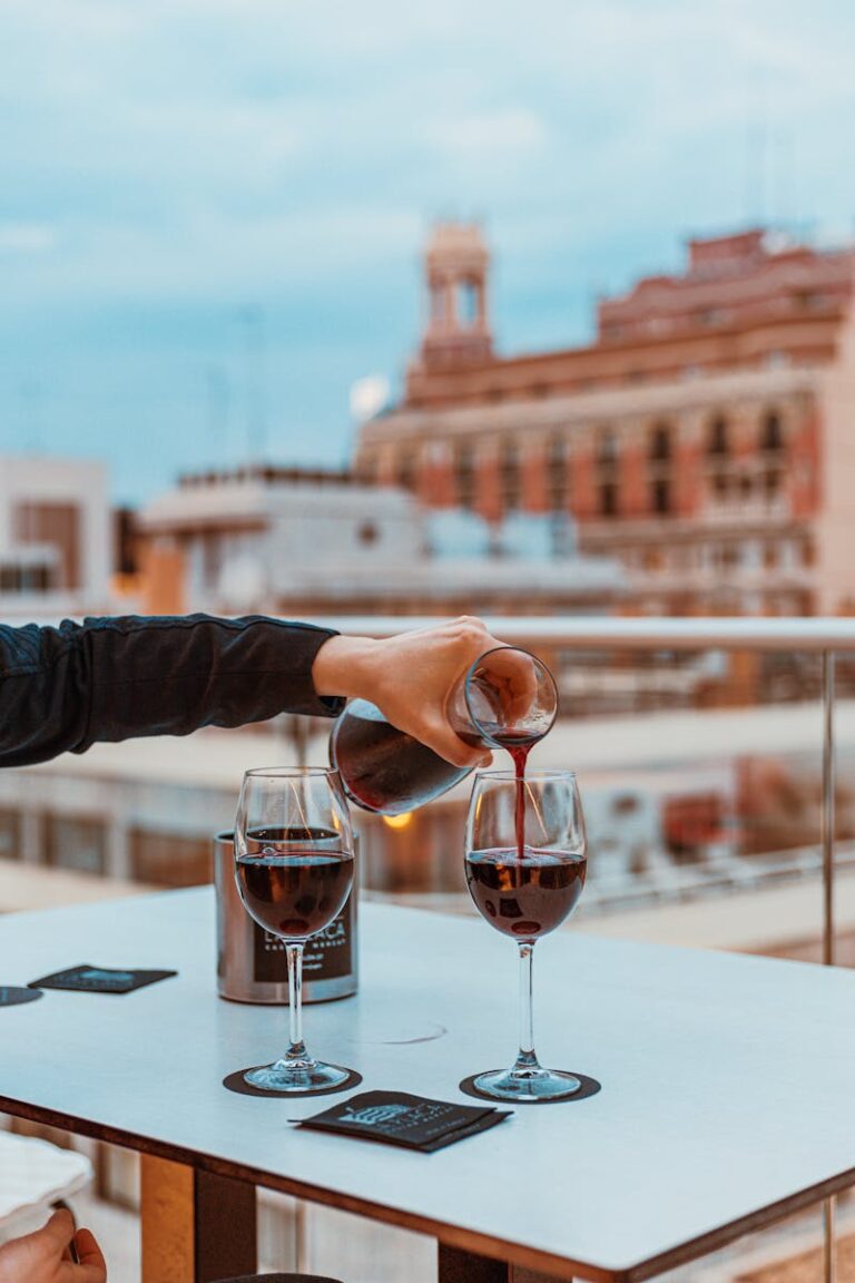 Een persoon schenkt rode wijn uit een karaf in twee glazen op een tafel. De tafel staat op een balkon met uitzicht op een stadsgezicht met historische gebouwen onder een blauwe lucht.