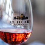 Close-up van een wijnglas met roséwijn met het logo "PURCARI" en de woorden "Fondat 1827 Premium Wine" erop gedrukt. Het glas maakt deel uit van een kalme en zacht verlichte setting, wat een warme en uitnodigende sfeer creëert.