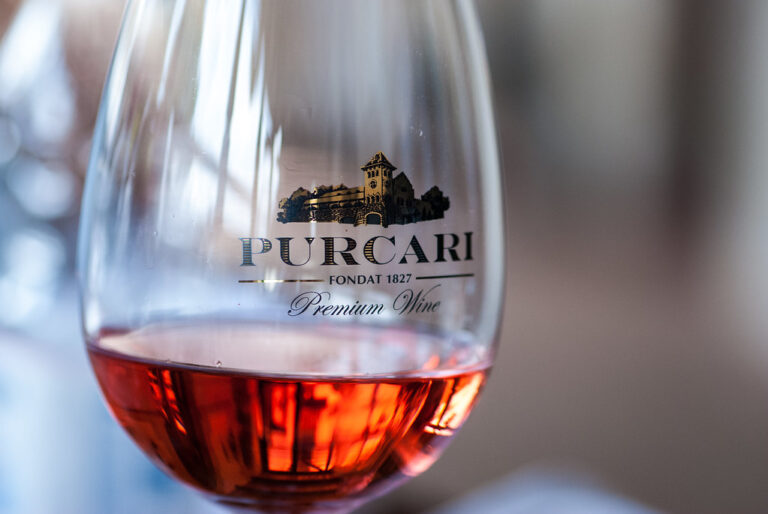 Close-up van een wijnglas met roséwijn met het logo "PURCARI" en de woorden "Fondat 1827 Premium Wine" erop gedrukt. Het glas maakt deel uit van een kalme en zacht verlichte setting, wat een warme en uitnodigende sfeer creëert.