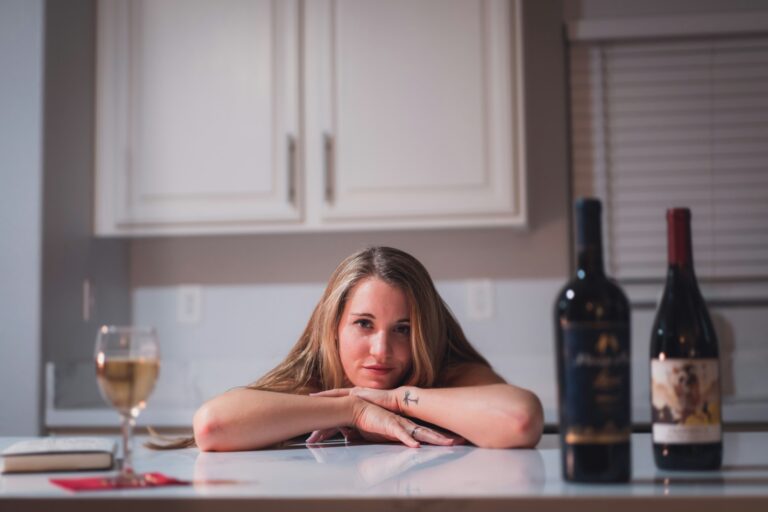 Een vrouw met lang haar laat haar armen en kin rusten op een aanrecht. Voor haar staan twee flessen wijn en een halfvol glas. De achtergrond bestaat uit witte kasten en een raam met jaloezieën.