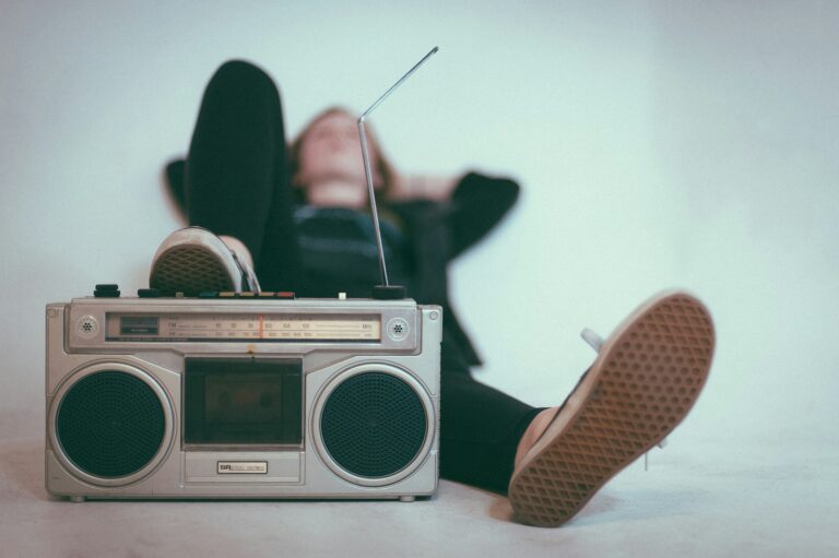 Een persoon ligt met gestrekte benen, gekleed in casual schoenen, voor een vintage boombox. De boombox heeft een antenne en cassettedeck, rustend op de grond. De achtergrond is effen en zacht gefocust.