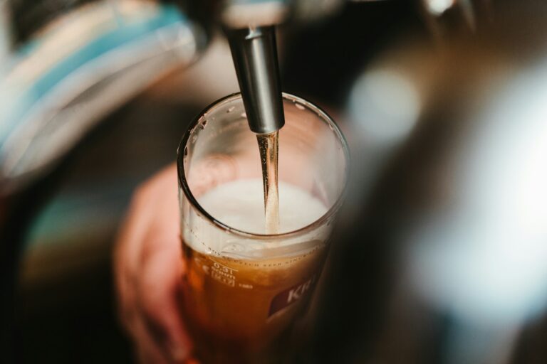 Een biertap schenkt een lichtbruin bier met een schuimkraag in een helder glas dat door een persoon wordt vastgehouden. De achtergrond is wazig.