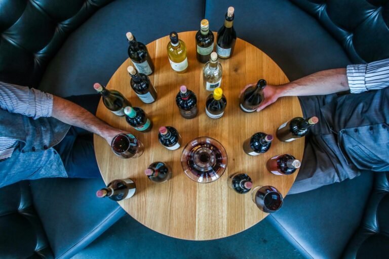 Een ronde houten tafel met verschillende flessen wijn in een cirkel. Twee personen, gedeeltelijk zichtbaar, zitten aan weerszijden, elk met een fles wijn. De tafel heeft een klein rond dienblad in het midden. De scène speelt zich af op een donkere bank.