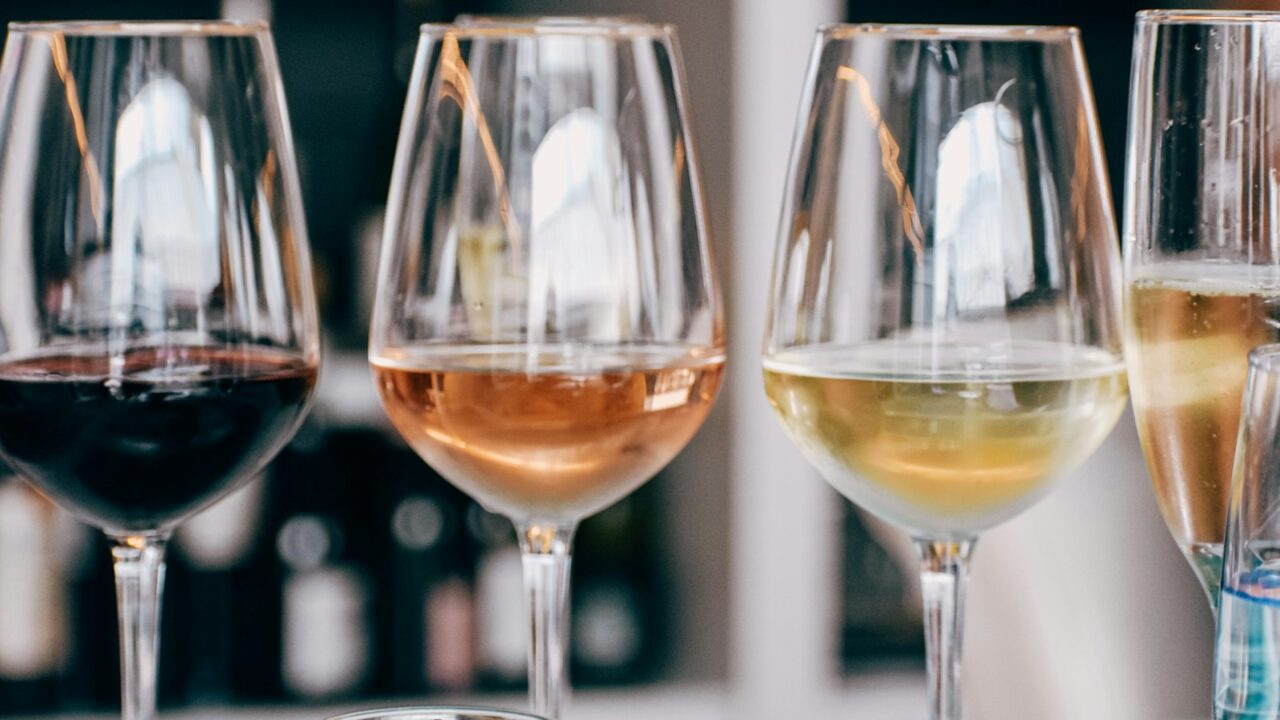 Vier wijnglazen gevuld met verschillende soorten wijn, waaronder rode, rosé, witte en mousserende wijn, staan op een tafel. De achtergrond toont onscherpe flessen op planken.