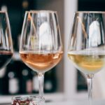 Vier wijnglazen gevuld met verschillende soorten wijn, waaronder rode, rosé, witte en mousserende wijn, staan op een tafel. De achtergrond toont onscherpe flessen op planken.