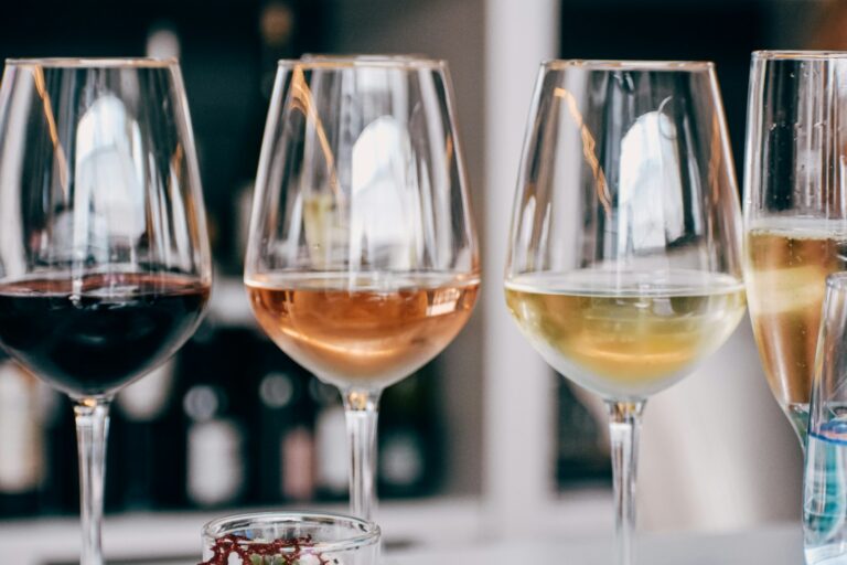 Vier wijnglazen gevuld met verschillende soorten wijn, waaronder rode, rosé, witte en mousserende wijn, staan op een tafel. De achtergrond toont onscherpe flessen op planken.