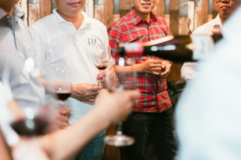 Een groep mensen staat bij elkaar met wijnglazen in de hand. Eén persoon schenkt wijn uit een fles. Ze zijn casual gekleed, één in een rood geruit overhemd en de ander in een witte polo. De achtergrond is voorzien van houten lambrisering.