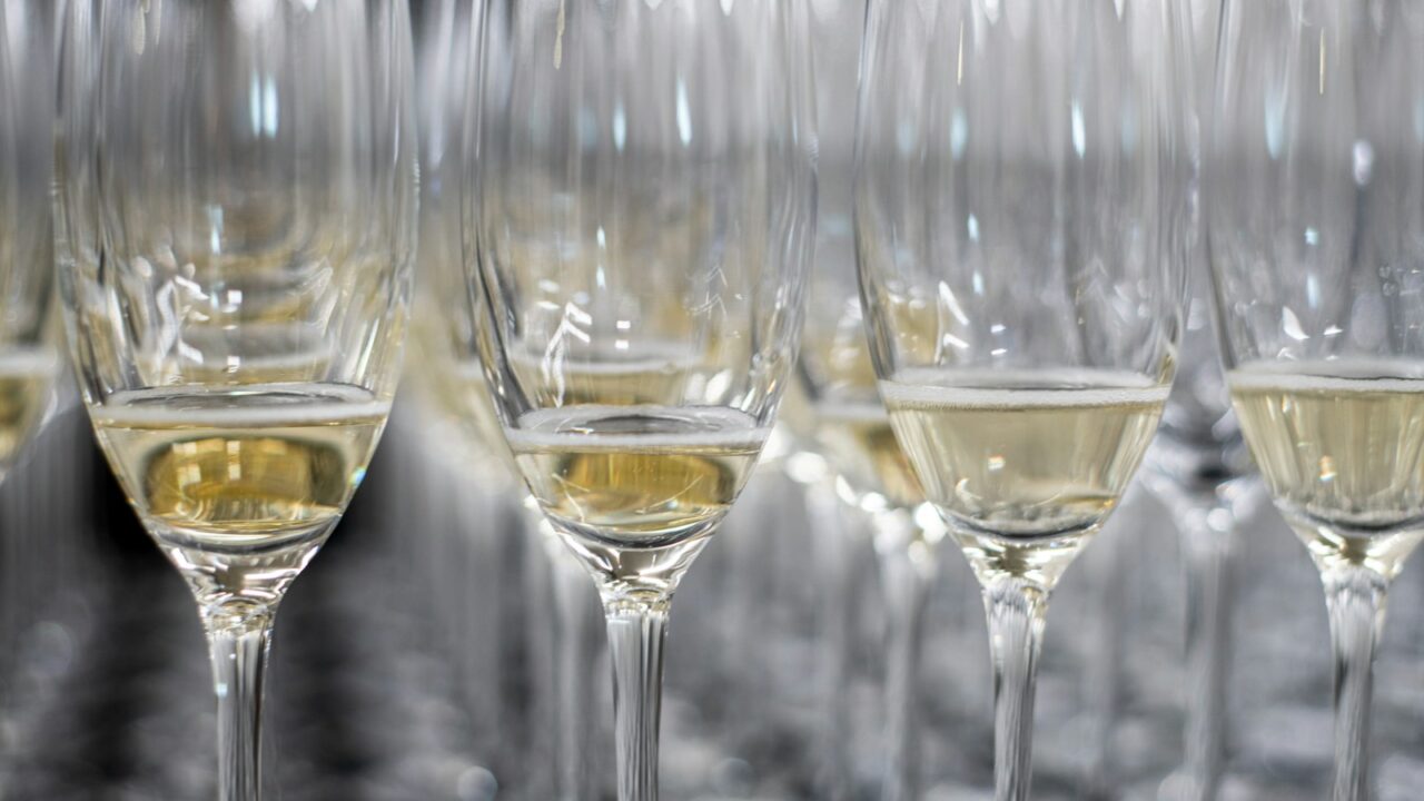 Rijen champagneglazen, gedeeltelijk gevuld met mousserende wijn, zijn gerangschikt op een reflecterend oppervlak. De glazen zijn dicht op elkaar geplaatst, waardoor een patroon ontstaat met de reflecties.