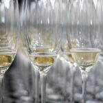 Rijen champagneglazen, gedeeltelijk gevuld met mousserende wijn, zijn gerangschikt op een reflecterend oppervlak. De glazen zijn dicht op elkaar geplaatst, waardoor een patroon ontstaat met de reflecties.