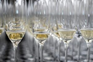 Rijen champagneglazen, gedeeltelijk gevuld met mousserende wijn, zijn gerangschikt op een reflecterend oppervlak. De glazen zijn dicht op elkaar geplaatst, waardoor een patroon ontstaat met de reflecties.