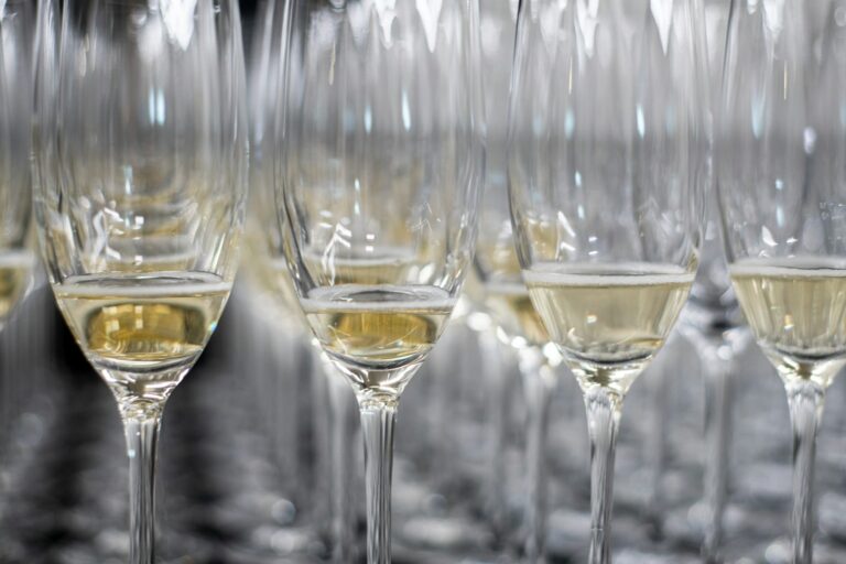 Rijen champagneglazen, gedeeltelijk gevuld met mousserende wijn, zijn gerangschikt op een reflecterend oppervlak. De glazen zijn dicht op elkaar geplaatst, waardoor een patroon ontstaat met de reflecties.