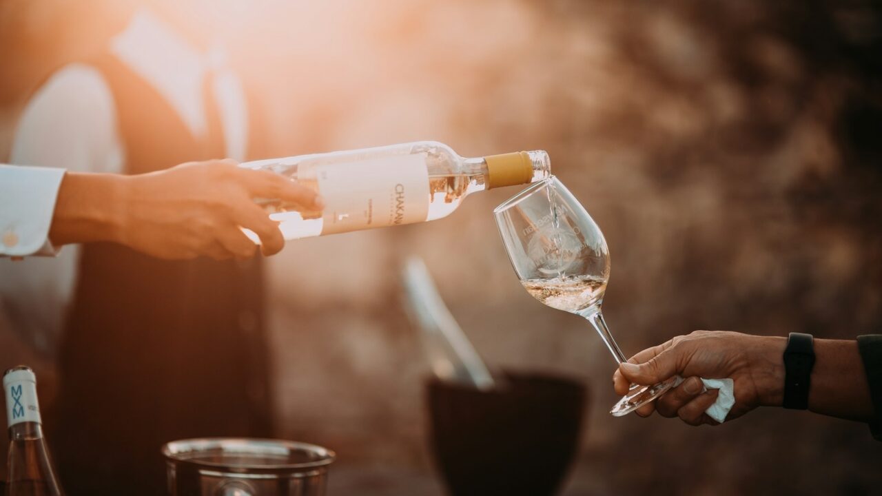 Een persoon giet witte wijn uit een fles in een wijnglas dat door een andere persoon wordt vastgehouden. De scène is warm verlicht, met een wazige achtergrond. Een ijsemmer met een fles staat op de achtergrond.