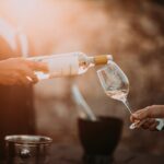 Een persoon giet witte wijn uit een fles in een wijnglas dat door een andere persoon wordt vastgehouden. De scène is warm verlicht, met een wazige achtergrond. Een ijsemmer met een fles staat op de achtergrond.
