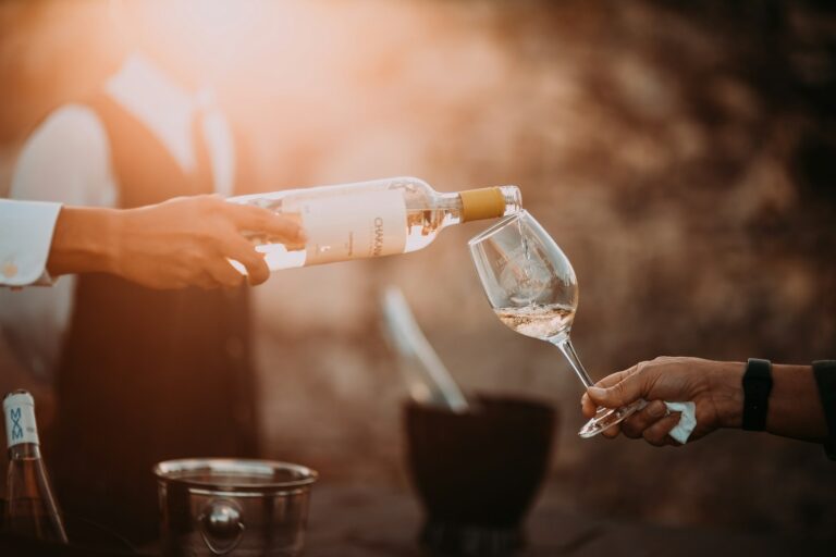 Een persoon giet witte wijn uit een fles in een wijnglas dat door een andere persoon wordt vastgehouden. De scène is warm verlicht, met een wazige achtergrond. Een ijsemmer met een fles staat op de achtergrond.