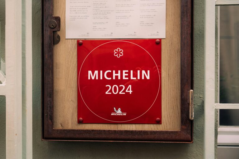 Een rode plaquette met het Michelin-sterlogo en het jaartal "2024" is op een houten oppervlak gemonteerd. Daarboven bevindt zich een ingelijst menu. De plaquette wordt begrensd door een grijze muur en een raamkozijn met een blauwgroene tint.