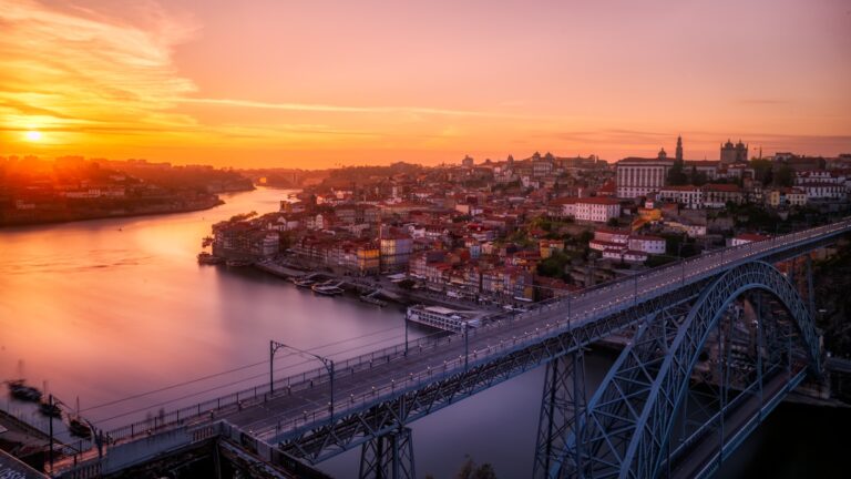 Een panoramisch uitzicht op een stadsgezicht tijdens zonsondergang, met een grote rivier die wordt doorsneden door een stalen boogbrug. De zon gaat onder aan de horizon, werpt een oranje en roze gloed over de lucht en reflecteert op het water. Stedelijke gebouwen staan langs de oevers van de rivier. Porto