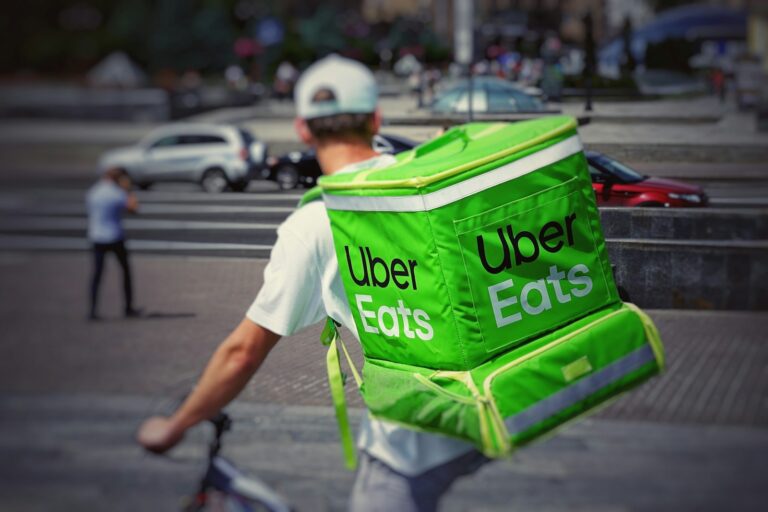Een persoon met een wit T-shirt en een pet fietst en draagt een grote groene rugzak met het logo van Uber Eats. De achtergrond toont een wazige stadsstraat met voetgangers en auto's.