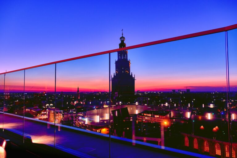 Uitzicht vanaf een dak bij zonsondergang over Groningen, met een silhouet van een de Martinitoren tegen een kleurrijke lucht van oranje, roze en blauw. Het stadsgezicht eronder is verlicht met straatlantaarns en een glazen barrière weerspiegelt het tafereel.