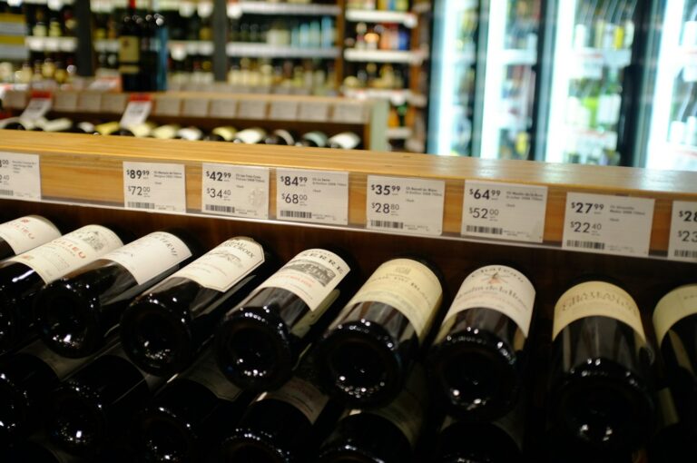 Wijnflessen staan op een plank in een winkel, elk met prijskaartjes erboven met verschillende prijzen en kortingen. Op de achtergrond is een koelgedeelte zichtbaar.