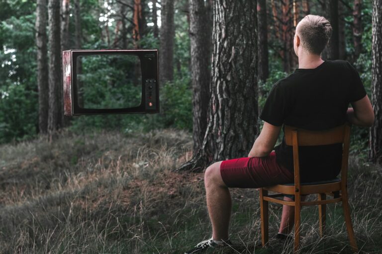 Een persoon zit op een houten stoel in een bos, met het gezicht naar een oude televisie die in de lucht hangt tussen de bomen. De scène combineert natuur en technologie op een ongewone manier.