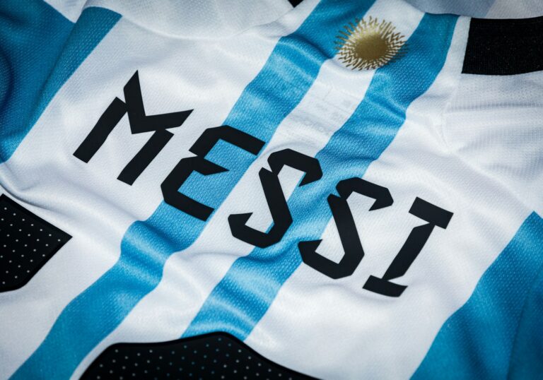 Close-up van een sportshirt met lichtblauwe en witte verticale strepen. De naam "Messi" is in vette zwarte letters gedrukt en er staat een klein gouden embleem met een zon erboven.