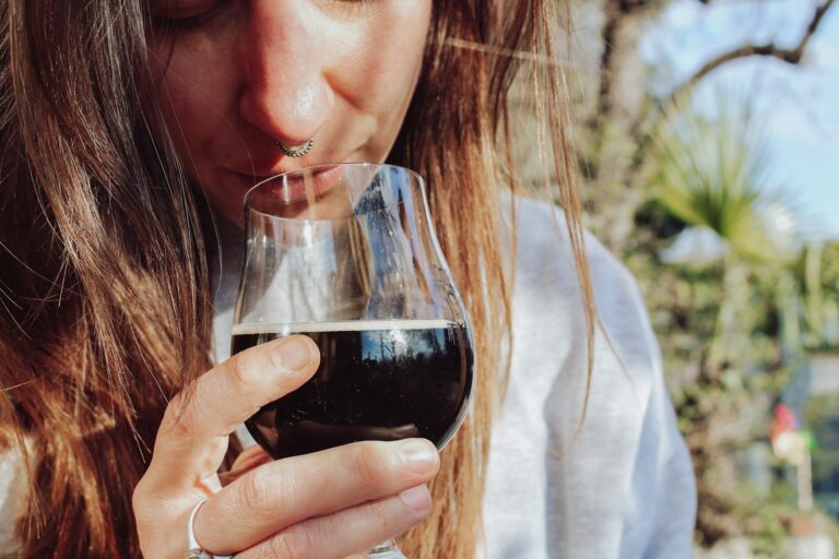 Een persoon met lang haar drinkt buiten een donker drankje uit een glas met steel. Het zonlicht versterkt het tafereel, met bomen die zachtjes vervagen op de achtergrond.
