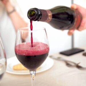 Een persoon schenkt rode wijn (Lambrusco) met een schuimende top in een wijnglas. Een tafelsetting met bestek en een bord met een broodstengel is op de achtergrond.