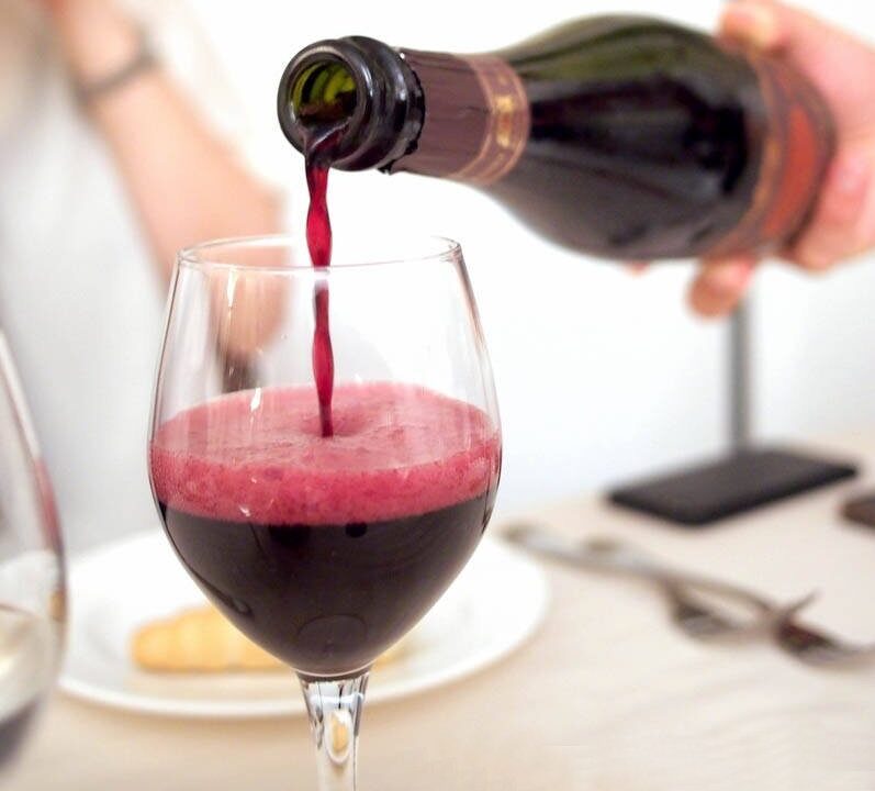 Een persoon schenkt rode wijn (Lambrusco) met een schuimende top in een wijnglas. Een tafelsetting met bestek en een bord met een broodstengel is op de achtergrond.