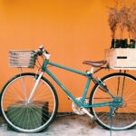 Een vintage-stijl blauwe fiets met een bruin zadel staat geparkeerd tegen een oranje muur. Er zit een mandje vooraan en een houten krat achterop met flessen en gedroogde planten. Een grote terracotta plantenbak met groen staat links.