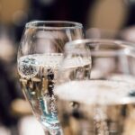 Close-up van twee glazen mousserende wijn gevuld met borrelende vloeistof. De glazen zijn geëtst met tekst die niet volledig leesbaar is. De achtergrond is zacht vervaagd, wat een warme en uitnodigende sfeer creëert.