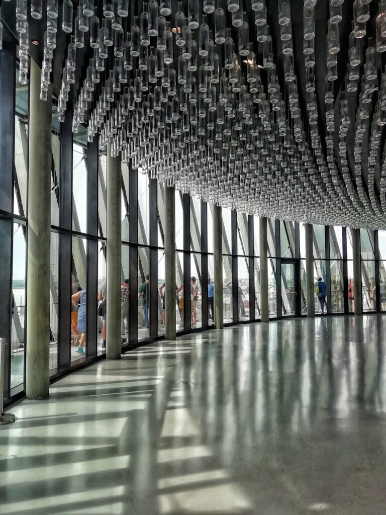 Cité du Vin. Een modern interieur met grote glazen ramen en een plafondinstallatie van talloze hangende glazen flessen. De vloer is van gepolijst beton en zonlicht stroomt erdoorheen, waardoor schaduwen en reflecties ontstaan. Mensen zijn buiten te zien door de ramen.