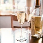 Een fles prosecco en twee gevulde champagneglazen staan op een houten tafel. Zonlicht stroomt door een raam naar binnen en creëert een heldere atmosfeer. Een wazig figuur is zichtbaar op de achtergrond.