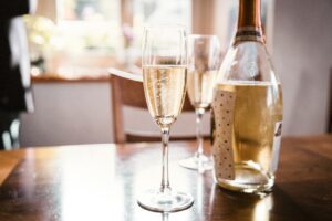 Een fles prosecco en twee gevulde champagneglazen staan op een houten tafel. Zonlicht stroomt door een raam naar binnen en creëert een heldere atmosfeer. Een wazig figuur is zichtbaar op de achtergrond.