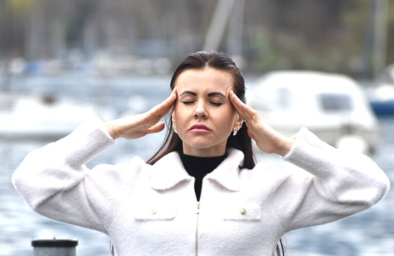 Hoofpijn. Een vrouw met lang haar en gesloten ogen staat buiten bij een watermassa en raakt haar slapen met beide handen aan. Ze draagt een witte jas en op de achtergrond zijn wazige boten zichtbaar.