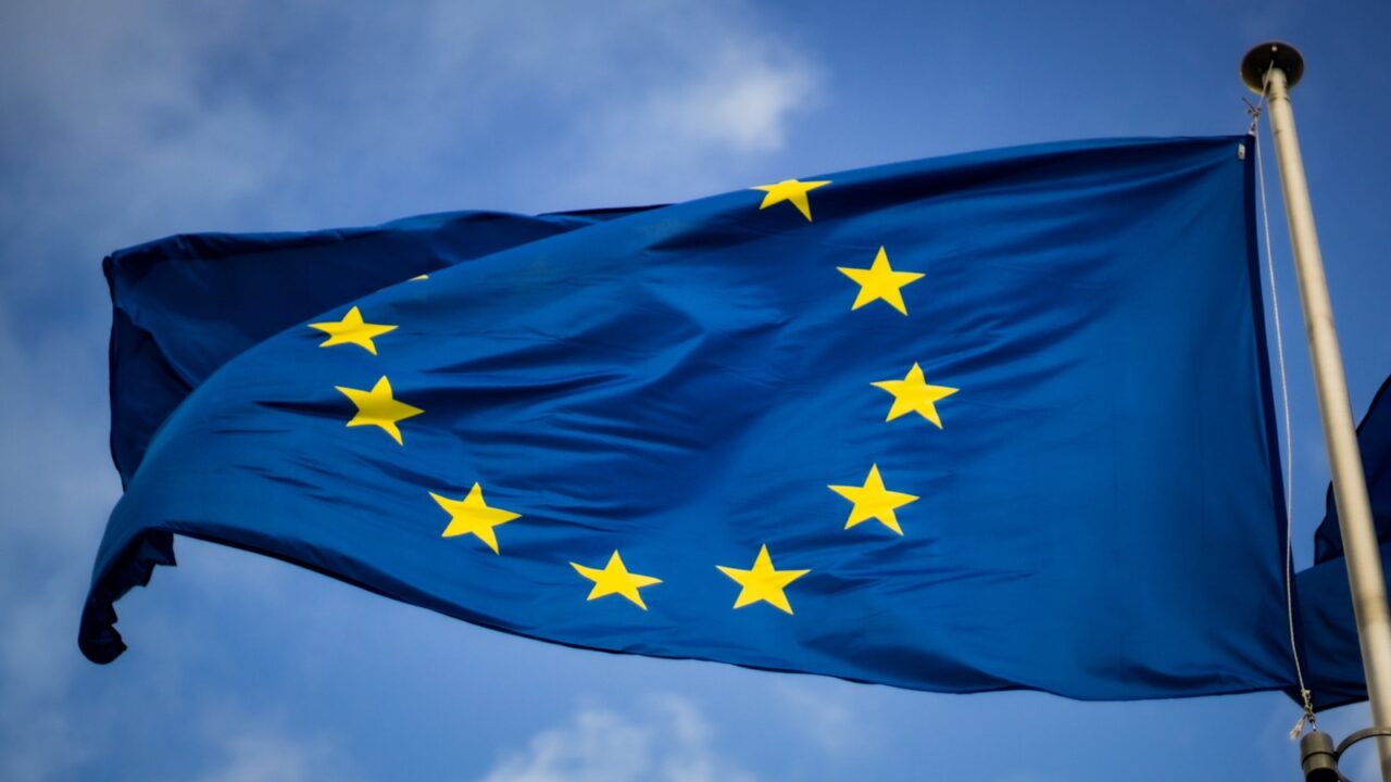 De afbeelding toont de vlag van de Europese Unie, met een cirkel van twaalf gouden sterren op een blauwe achtergrond, wapperend tegen een gedeeltelijk bewolkte lucht.