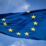 De afbeelding toont de vlag van de Europese Unie, met een cirkel van twaalf gouden sterren op een blauwe achtergrond, wapperend tegen een gedeeltelijk bewolkte lucht.