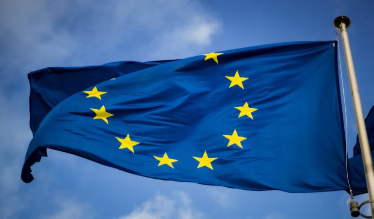 De afbeelding toont de vlag van de Europese Unie, met een cirkel van twaalf gouden sterren op een blauwe achtergrond, wapperend tegen een gedeeltelijk bewolkte lucht.