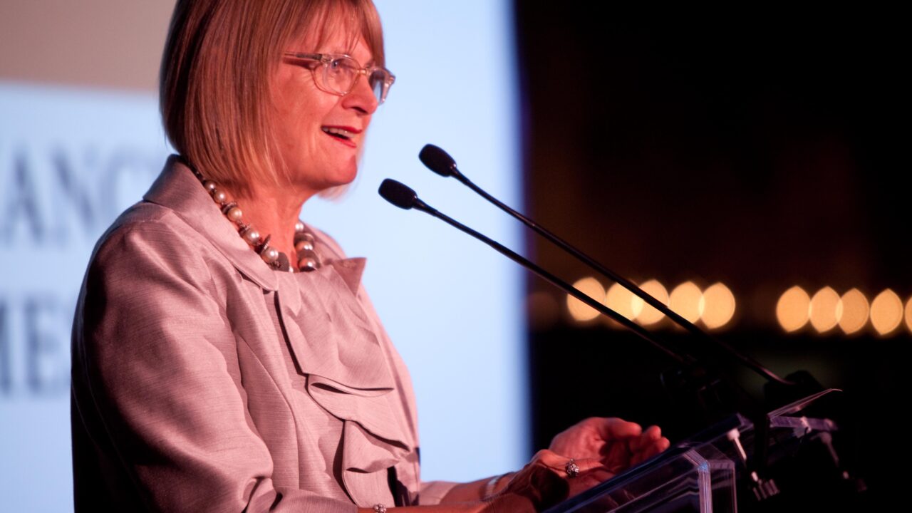 Een vrouw met een bril en een lichtgekleurd pak spreekt op een podium met twee microfoons, lichtjes glimlachend. De achtergrond is wazig met wat lichtjes zichtbaar. Jancis Robinson