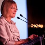 Een vrouw met een bril en een lichtgekleurd pak spreekt op een podium met twee microfoons, lichtjes glimlachend. De achtergrond is wazig met wat lichtjes zichtbaar. Jancis Robinson