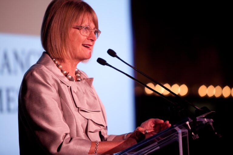 Een vrouw met een bril en een lichtgekleurd pak spreekt op een podium met twee microfoons, lichtjes glimlachend. De achtergrond is wazig met wat lichtjes zichtbaar. Jancis Robinson