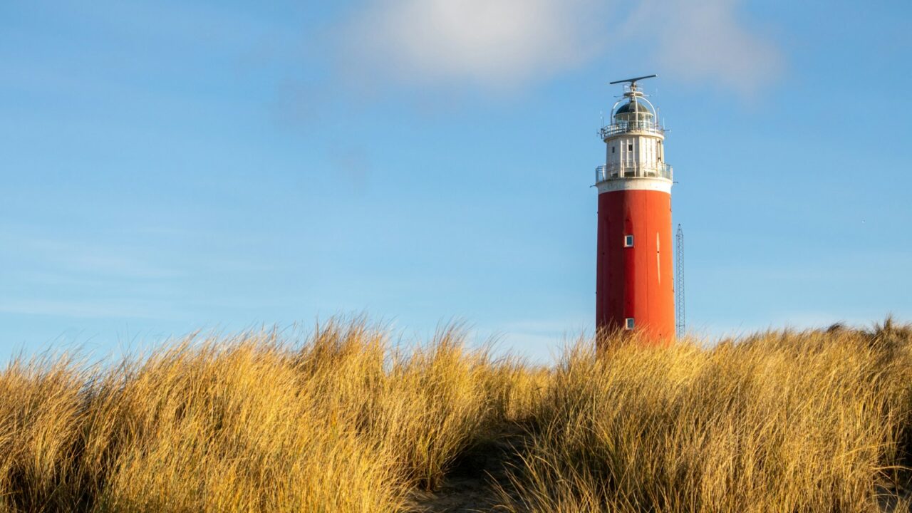 Een rode vuurtoren met een witte top staat achter een veld met hoog, goudkleurig gras onder een blauwe lucht met een paar wolken. Texel