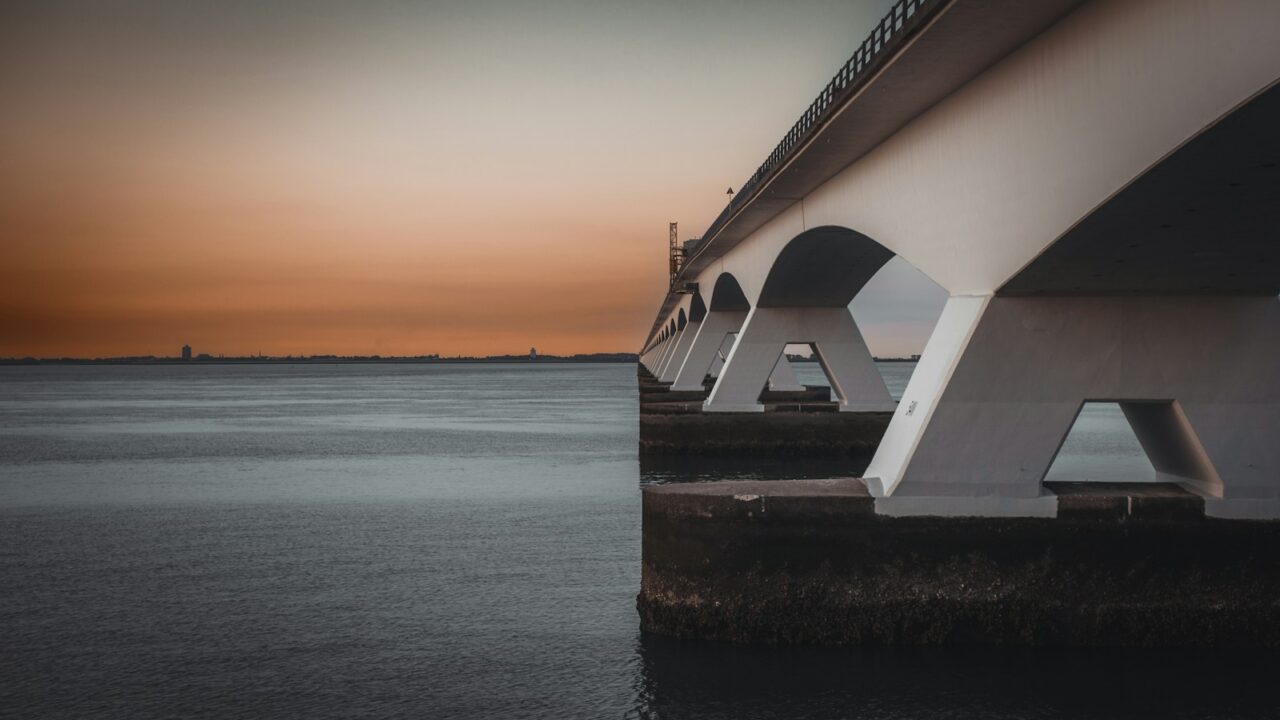 Een grote betonnen brug strekt zich uit over een kalm waterlichaam, met zijn bogen die in het water weerspiegelen. De lucht is een gradiënt van oranje en grijs, wat duidt op zonsondergang of zonsopgang. De horizon toont verre gebouwen en een vlakke kustlijn. Zeeland