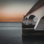 Een grote betonnen brug strekt zich uit over een kalm waterlichaam, met zijn bogen die in het water weerspiegelen. De lucht is een gradiënt van oranje en grijs, wat duidt op zonsondergang of zonsopgang. De horizon toont verre gebouwen en een vlakke kustlijn. Zeeland