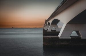 Een grote betonnen brug strekt zich uit over een kalm waterlichaam, met zijn bogen die in het water weerspiegelen. De lucht is een gradiënt van oranje en grijs, wat duidt op zonsondergang of zonsopgang. De horizon toont verre gebouwen en een vlakke kustlijn. Zeeland
