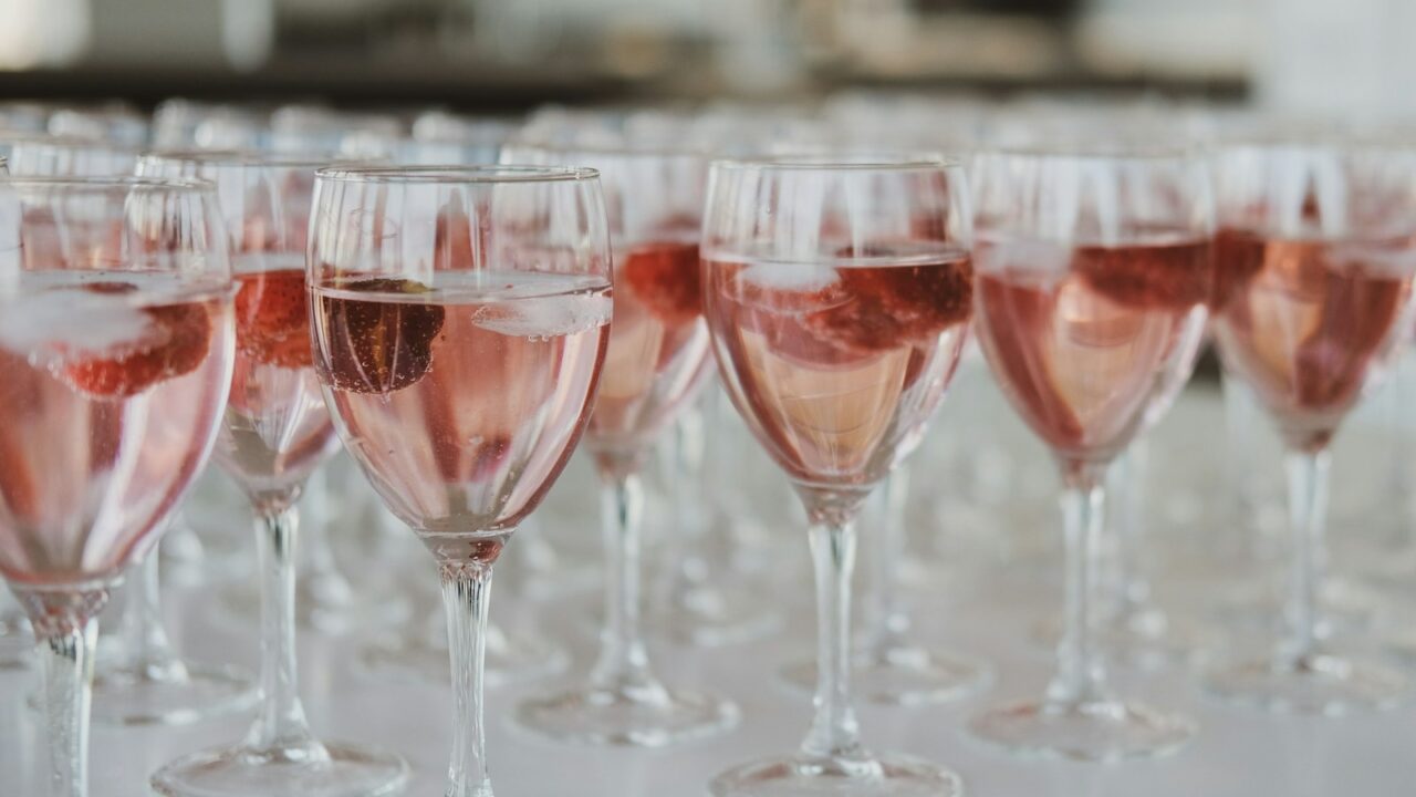 Verschillende wijnglazen gevuld met een roze drankje en ijsblokjes staan in rijen op een wit oppervlak. Elk glas bevat een stuk fruit, mogelijk een aardbei. De achtergrond is onscherp.