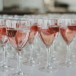 Verschillende wijnglazen gevuld met een roze drankje en ijsblokjes staan in rijen op een wit oppervlak. Elk glas bevat een stuk fruit, mogelijk een aardbei. De achtergrond is onscherp.