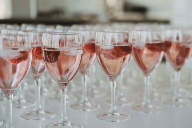 Verschillende wijnglazen gevuld met een roze drankje en ijsblokjes staan in rijen op een wit oppervlak. Elk glas bevat een stuk fruit, mogelijk een aardbei. De achtergrond is onscherp.