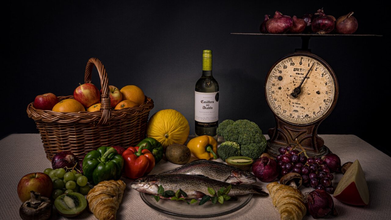 Een stilleven met een mand appels, een hele vis op een glazen bord, een fles wijn, diverse soorten fruit en groenten, een punt kaas, een oude weegschaal en een bos rode uien op een donkere achtergrond.