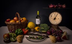 Een stilleven met een mand appels, een hele vis op een glazen bord, een fles wijn, diverse soorten fruit en groenten, een punt kaas, een oude weegschaal en een bos rode uien op een donkere achtergrond.